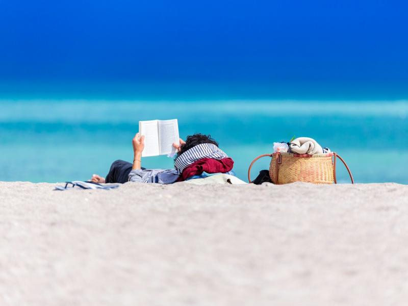 Person på strand der ligger på ryggen mens de læser en bog